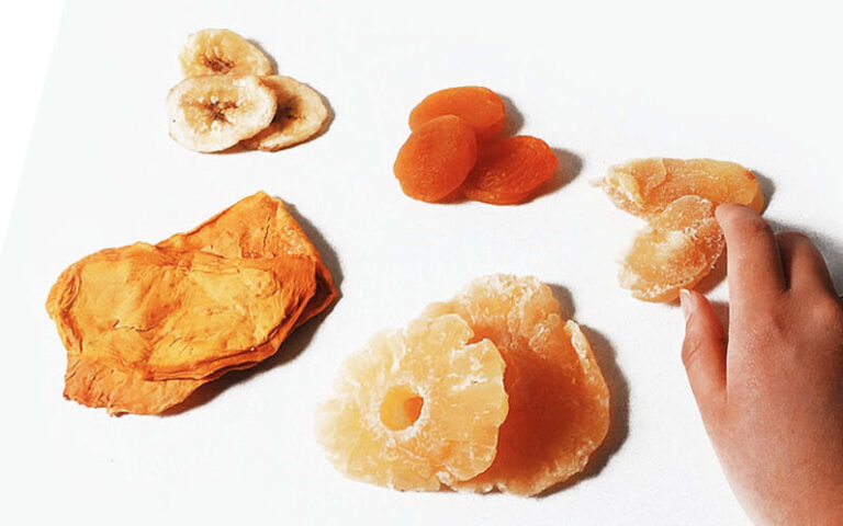 Dried fruit/plant-powered treats on off-white surface. Hand grabbing dried peaches
